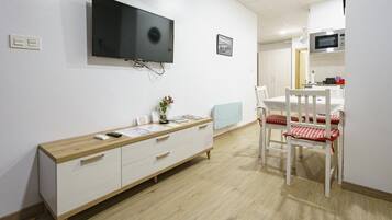 A room with a TV mounted on the wall, a wooden cabinet, a dining table with chairs, and a kitchen area in the background.