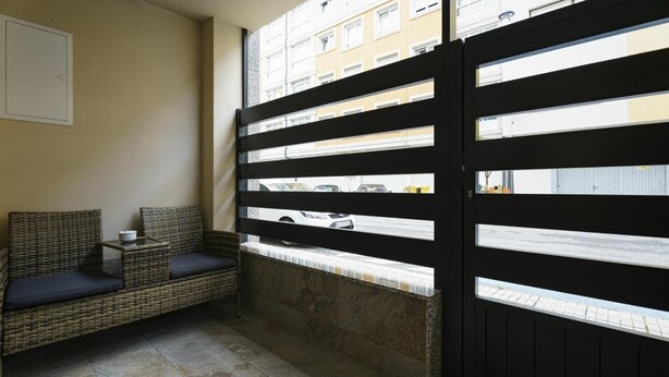 Une pièce avec un canapé en osier, une petite table et une vue par une fenêtre ornée d’une grille en métal noir.