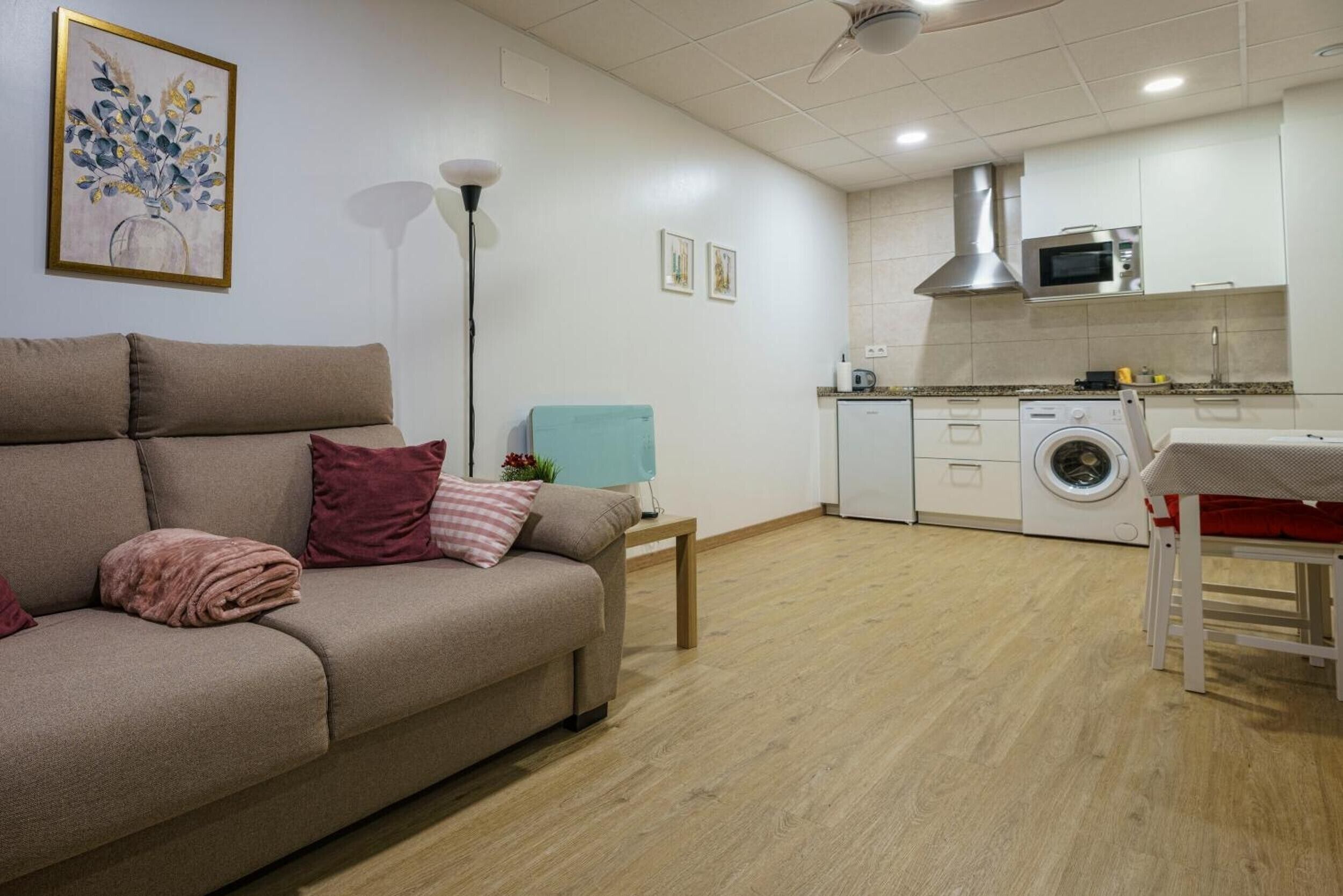 Una sala de estar con sofá, mesa, lavadora y una zona de cocina con armarios y microondas.