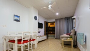 Un comedor con una mesa y sillas, un ventilador de techo, un reloj, un televisor, un sofá, una mesita con una lámpara y una ventana con cortinas.