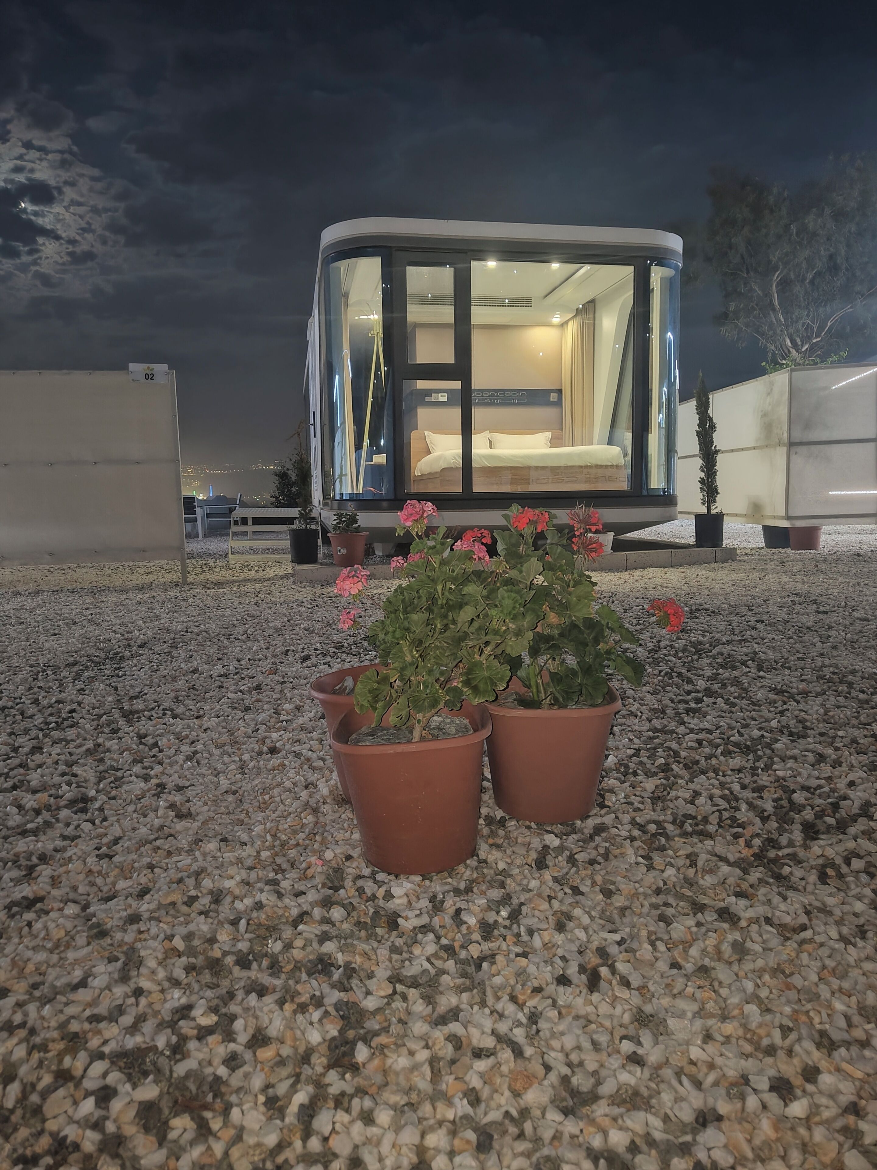 Una habitación moderna con paredes de vidrio, una cama y un ventanal, rodeada de grava y plantas en macetas.