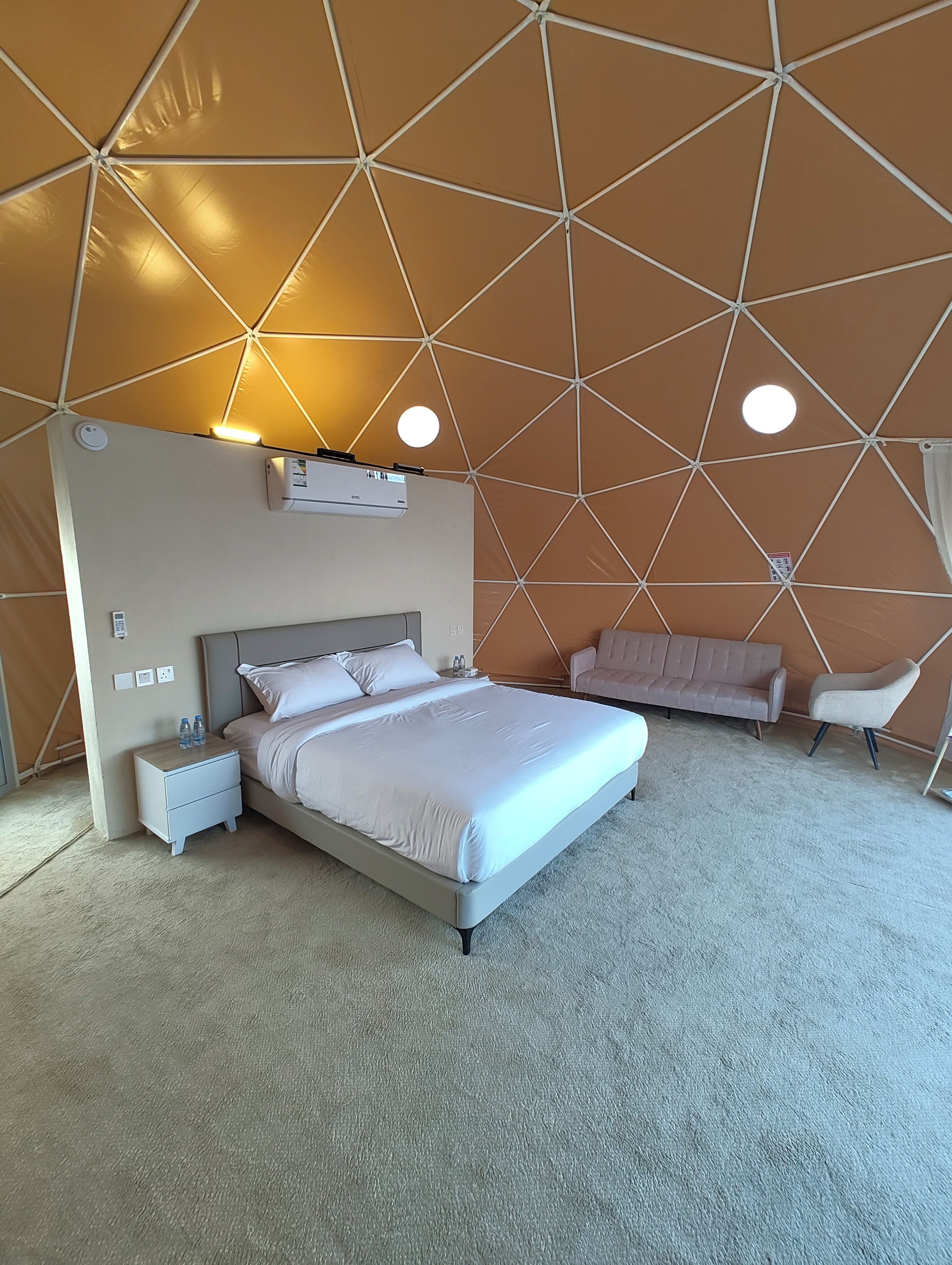 Un dormitorio moderno con techo de estampado geométrico, una cama grande con ropa blanca, una mesita de noche, un sofá y una silla.