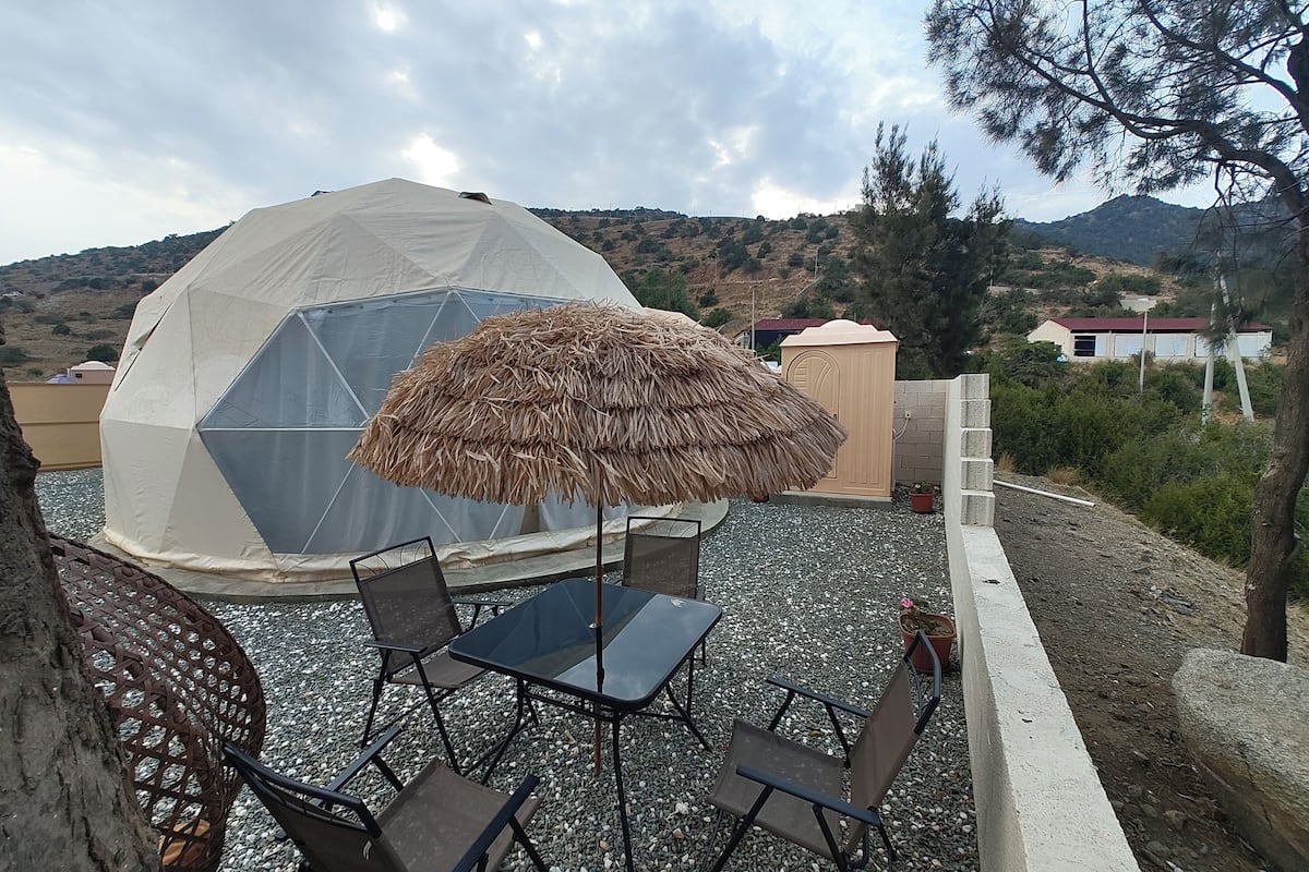 A geodesic dome structure with a thatched umbrella, surrounded by a gravel patio with metal chairs and a table.