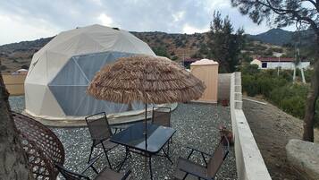 A geodesic dome structure with a thatched umbrella, surrounded by a gravel patio with metal chairs and a table.