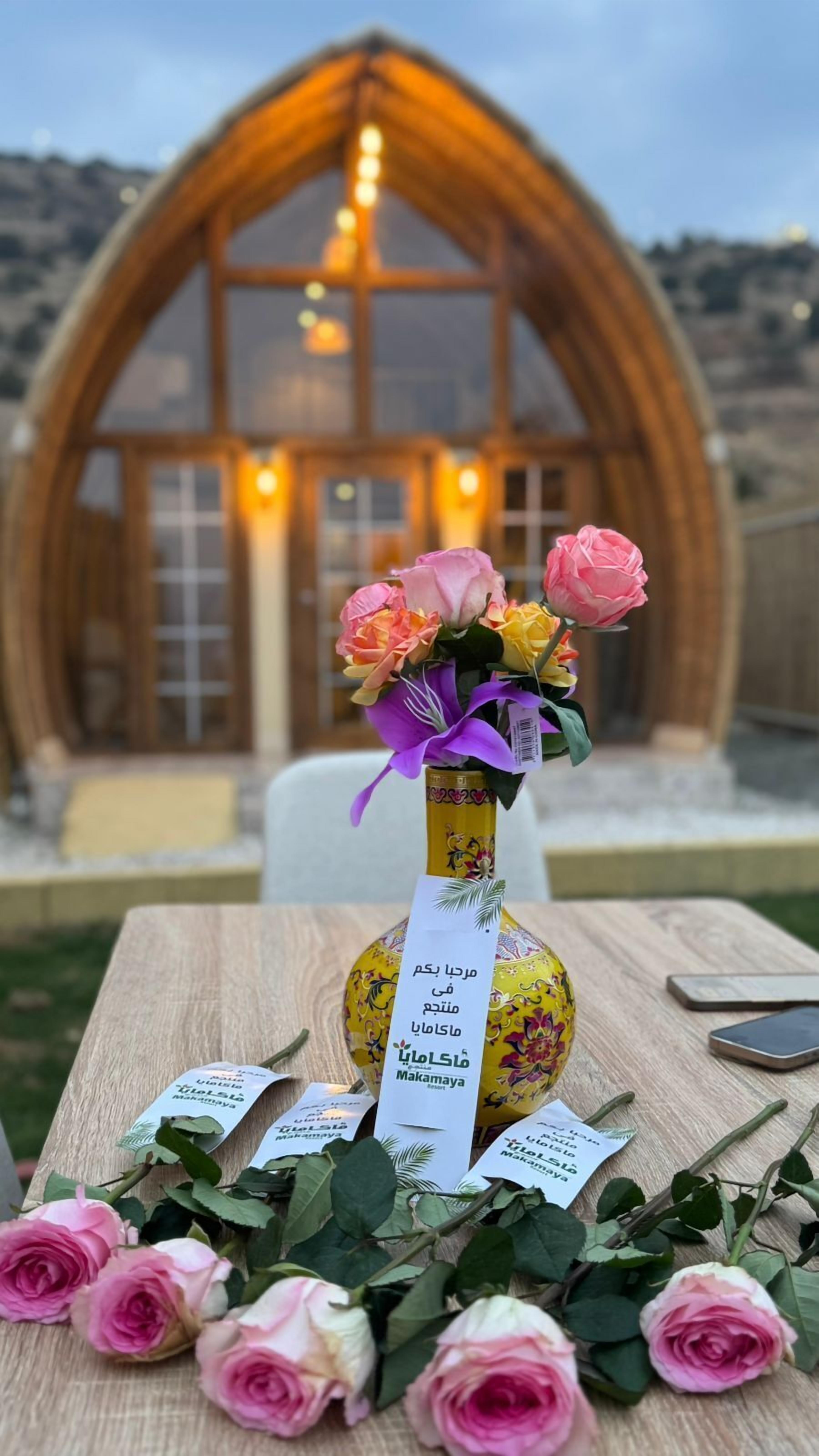 Una mesa con una botella de vino Makamaya, un ramo de rosas y una mesa de madera con vista a un edificio al fondo.
