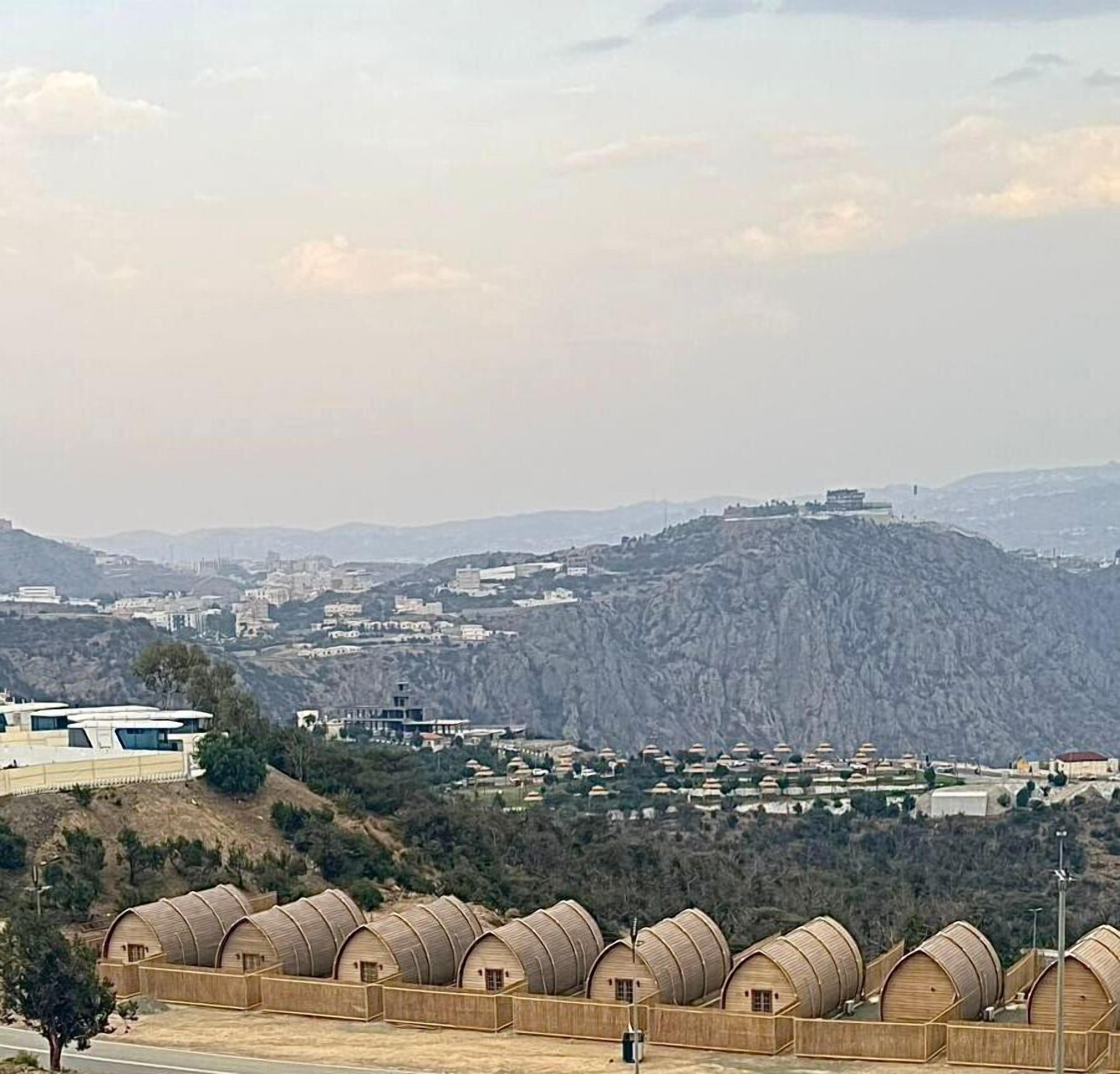 A view of a mountainous area with a series of wooden barrel-shaped buildings in the foreground, a distant cityscape, and a prominent hill with structures on top.