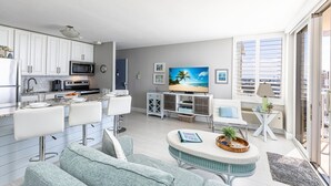 A modern kitchen with white cabinets, a microwave, and a dining area with bar stools. There is a TV in the living room with a beach painting on the wall. - Estero Beach Tennis 1004c Direct Gulf Water Views (Fort Myers Beach)