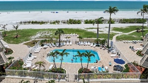 A pool area with lounge chairs and palm trees, overlooking a beach with people and boats.
