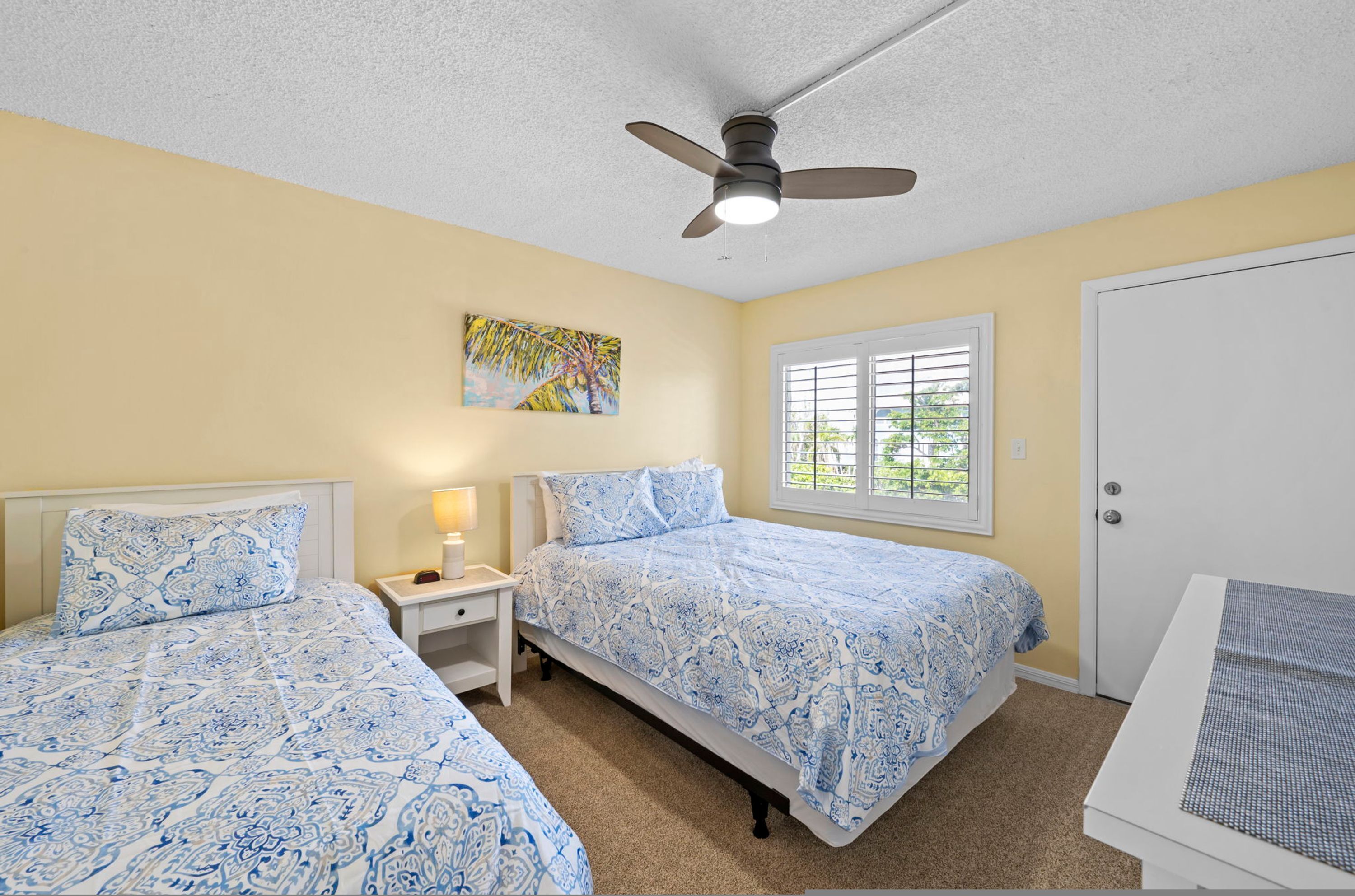 A bedroom with two beds, a ceiling fan, a window with blinds, and a door.