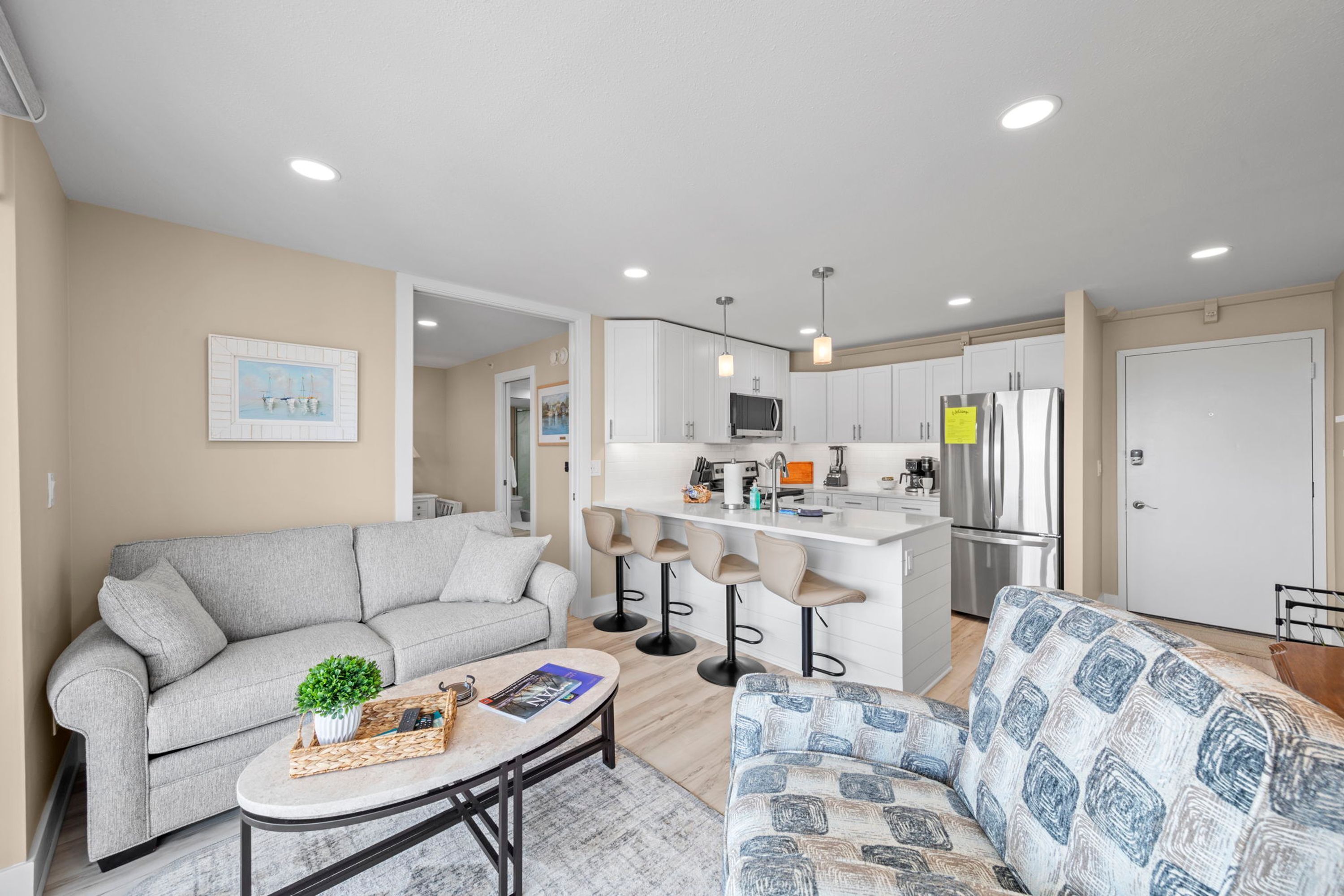 A modern living room with a grey sofa, a patterned sofa, and a coffee table. There is a kitchen area with white cabinets and a stainless steel refrigerator.