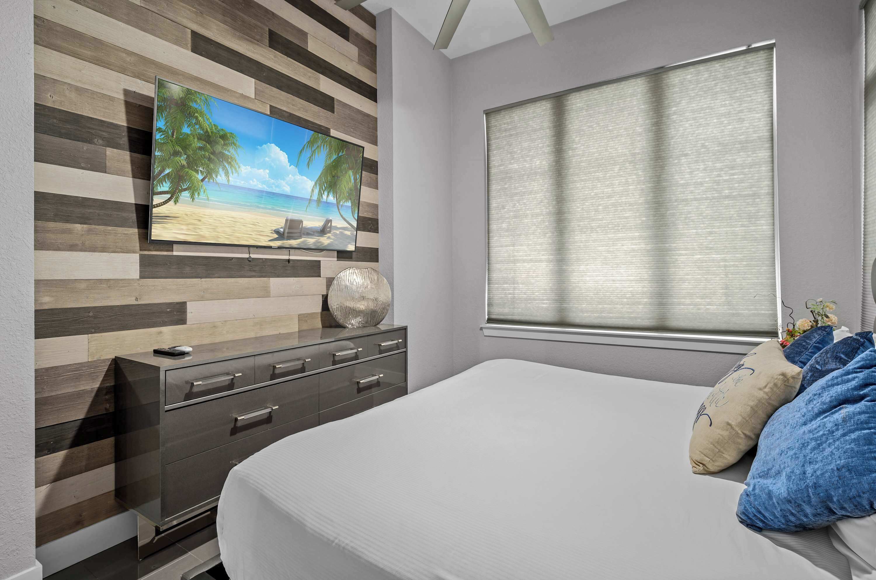 Une chambre à coucher comprenant un lit, une commode, un téléviseur fixé au mur (qui est en bois) et une fenêtre avec des stores.