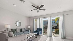 A modern living room with a grey sofa, a blue armchair, and a coffee table. There is a ceiling fan, a round mirror, and a view of the outside through large glass doors.