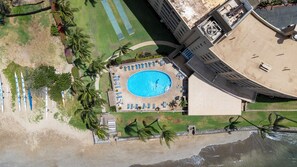 Eine Vogelperspektive zeigt einen Urlaubsort mit einem zentral gelegenen Pool, umgeben von Liegestühlen, Palmen und einem Strandabschnitt mit Kajaks.
