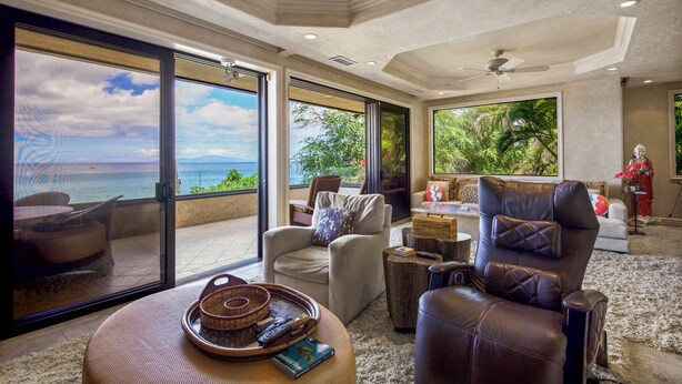 Uma sala de estar espaçosa com vista para o oceano, uma varanda com mesa e cadeiras, e um espaço de estar com poltrona de couro e sofá.