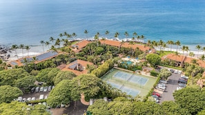 An aerial view of a resort with a beach, palm trees, solar panels on buildings, a tennis court, and a parking area.