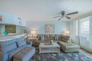 A living room with a sofa, coffee table, and a ceiling fan. There is a clock on the wall and a window with blinds. - 402 Southpoint in Ponce Inlet, FL (Ponce Inlet)