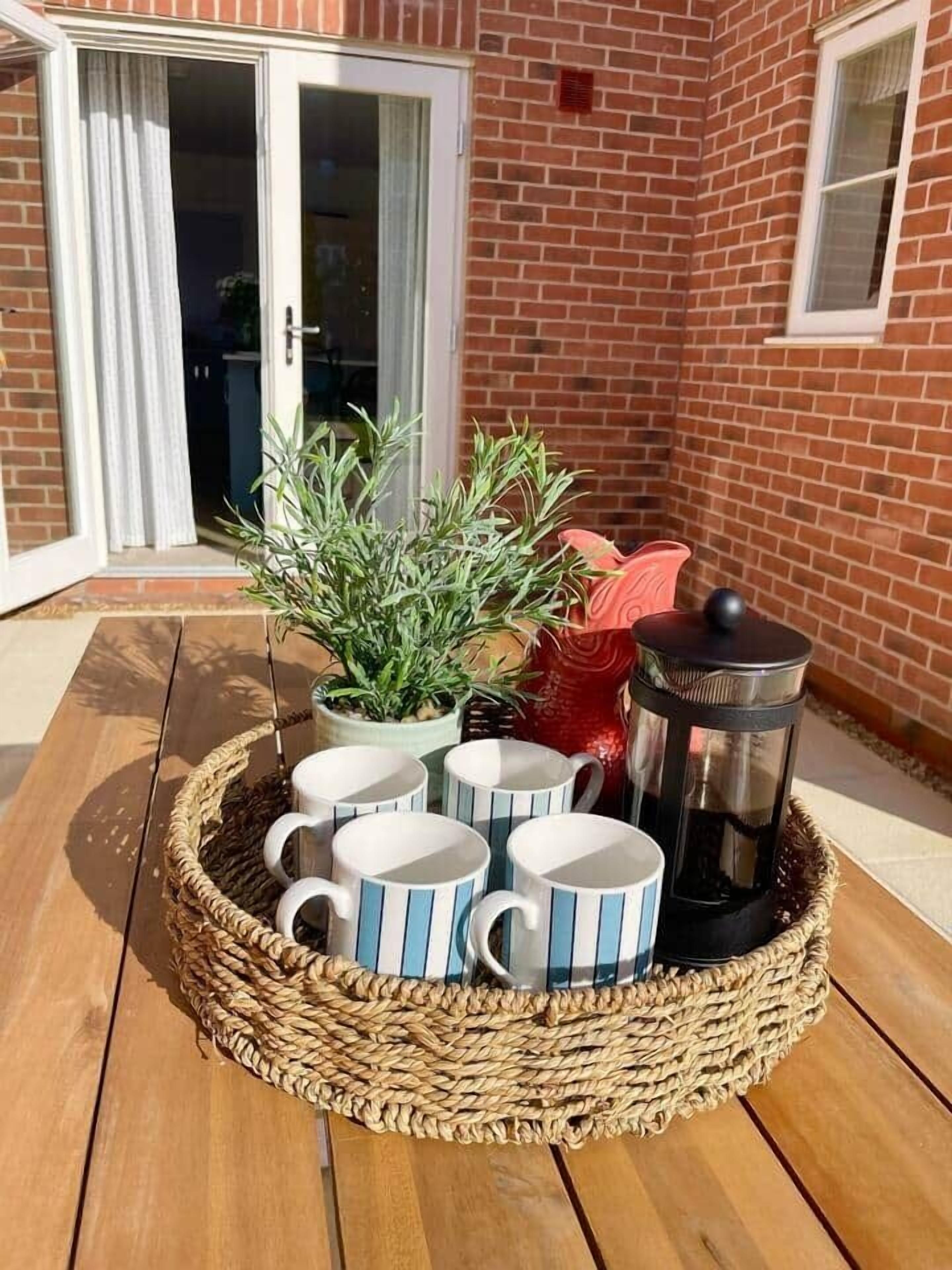 Una bandeja de mimbre sobre una cubierta de madera, con tazas a rayas, una termo y una planta en maceta.