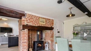 A cozy living room with a brick fireplace, a dining area with chairs, and a kitchen in the background. - West Cottage. Central, spacious, 2 mins to beach. (Cromer)
