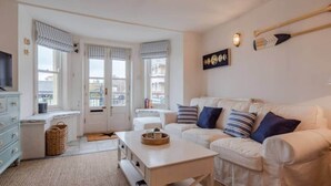 A cozy living room with a white sofa, a small table, and a television. There are windows with striped curtains, a wall-mounted light, and a framed picture.