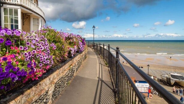 A coastal walkway with a stone wall and flowering plants, leading to a beach with a boat and a sign for 'Sea Cream'.