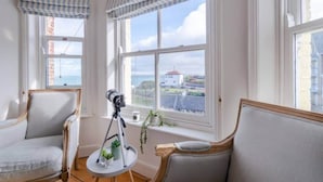A room with two armchairs, a tripod with a camera, and a view of the sea and buildings through large windows with striped valances.