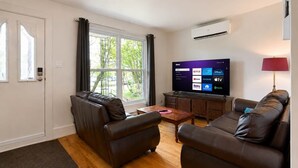 Sala de estar con sofás de cuero, una mesa de centro de madera, un televisor de pantalla plana mostrando servicios de transmisión y una ventana con cortinas.