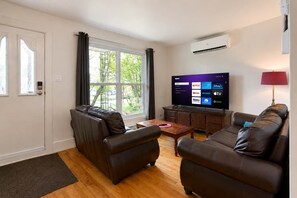 A living room with leather sofas, a wooden coffee table, a flat-screen TV displaying streaming services, and a window with curtains.