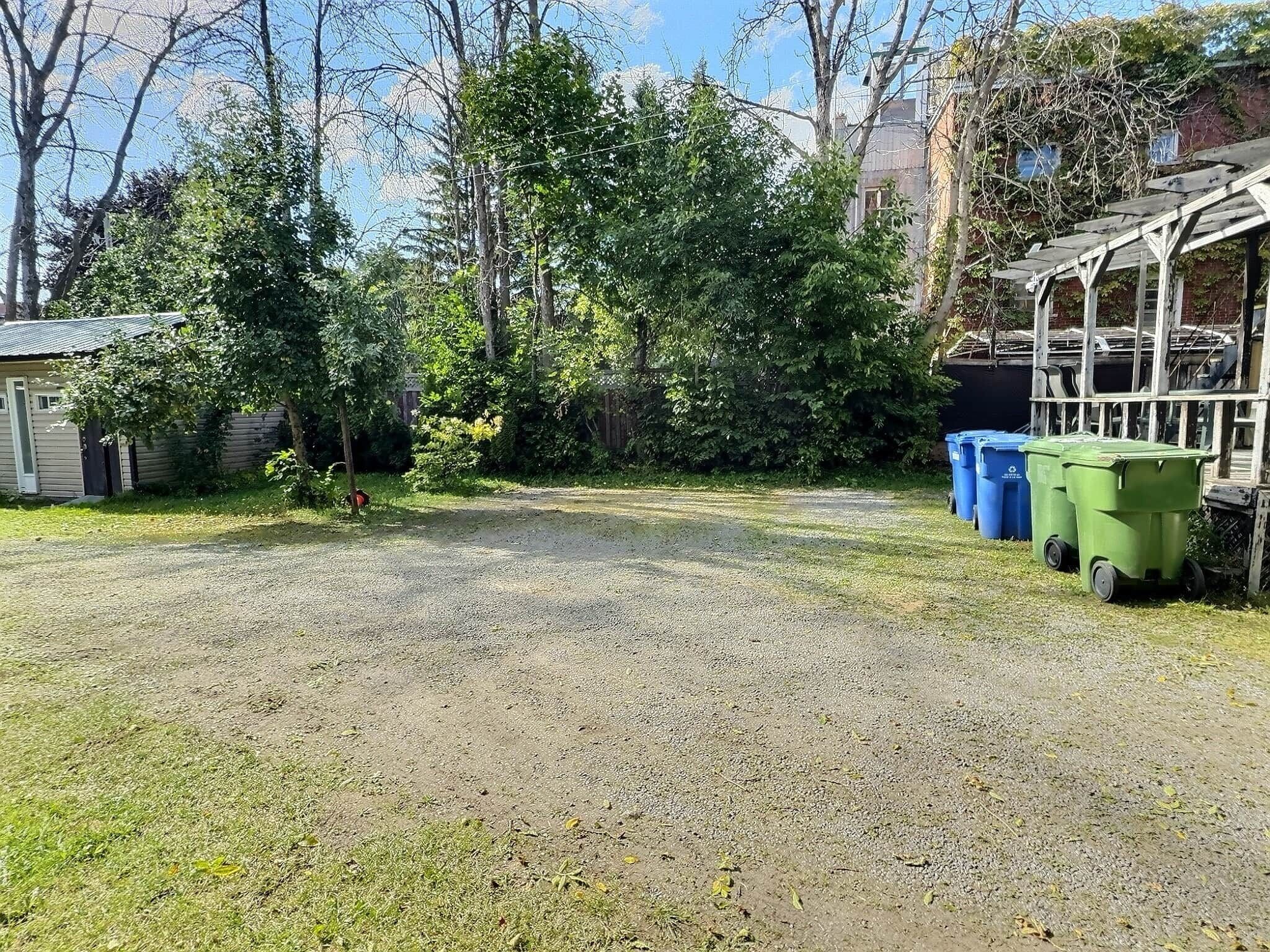 A gravel yard with a small shed, a tree, and a covered structure with blue and green bins.