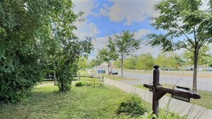A grassy area with a wooden signpost, a paved path, and a road with cars in the background. - Chez Claude appartement millésimé-free parking (Québec City)
