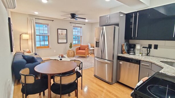 A modern kitchen with stainless steel appliances, a dining area with a round table and chairs, and a living space with a sofa and a ceiling fan. - Spacious 1BD With Balcony (Philadelphia)