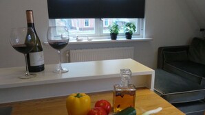 Eine Küche mit Holztisch, einer Weinflasche und einem Glas auf der Arbeitsplatte. Auf dem Tisch liegen Gemüse und ein Fenster mit Blick auf Gebäude.