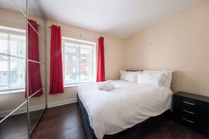 Ein Schlafzimmer mit einem Bett, einem Nachttisch, einem Fenster mit Jalousien und roten Vorhängen.
