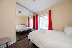 A bedroom with a large mirror, a bed with white linens, red curtains, and a bench.
