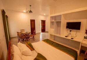 A hotel room with a bed, a TV mounted on the wall, a dining area with chairs, and a wooden door. - Shambala Resort (Bommayapalayam)