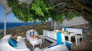 Un espace extérieur abrité, aménagé avec des meubles en osier, une table dressée avec des assiettes et des tasses, et une vue sur la mer.