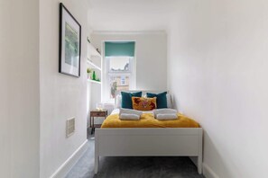 A narrow hallway leading to a bed with a yellow blanket, teal pillows, and a floral pillow.