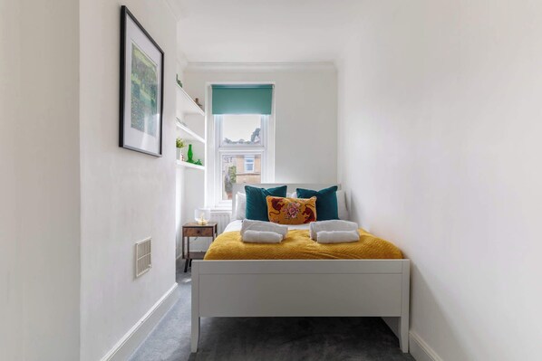 Un couloir étroit mène à un lit recouvert d’une couverture jaune, agrémenté d’oreillers turquoise et d’un coussin à motifs floraux.