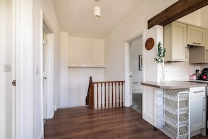 A hallway with wooden flooring, a wooden railing, a kitchen area with white cabinets, and a small plant on the counter.