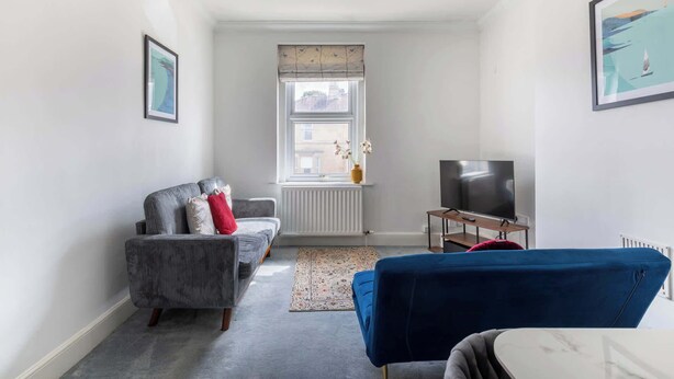 A compact living space with a sofa, a blue chair, a TV, and a radiator. - Refurbished Modern 2BD Flat, Lower Oldfield Park (Bath and North East Somerset)