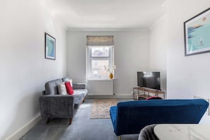 A compact living space with a sofa, a blue chair, a TV, and a radiator. - Refurbished Modern 2BD Flat, Lower Oldfield Park (Bath and North East Somerset)