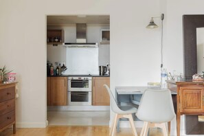 A compact kitchen with wooden cabinets, a built-in oven, and a dining area with a table and chairs. - Eclectically Curated 2BD Flat in Canary Wharf (Greater London)