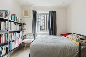 A bedroom with a bed, a bookshelf, a chair, and a window with curtains. - Eclectically Curated 2BD Flat in Canary Wharf (Greater London)