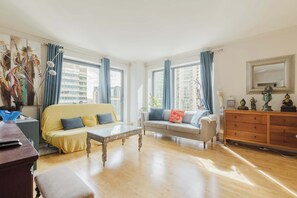 A spacious living room with a yellow sofa, a grey sofa, and a wooden cabinet. Large windows with blue curtains allow natural light to fill the room. - Eclectically Curated 2BD Flat in Canary Wharf (Greater London)