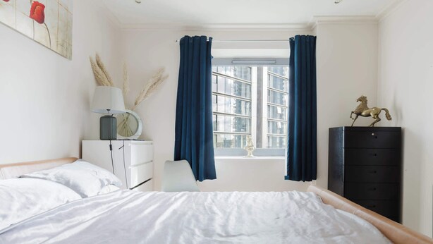 Ein Schlafzimmer mit Bett, Nachttisch, Lampe, Kommode und einem Fenster mit Vorhängen.
