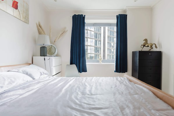 A bedroom with a bed, a nightstand, a lamp, a dresser, and a window with curtains.