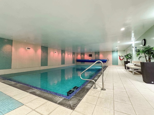 An indoor swimming pool with a blue tiled floor, a metal pool ladder, and a bench along the wall.