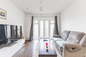 A living room with a sofa, a coffee table with a flower, a TV, and a window with curtains. - Modern two-bedroom flat, fully equipped (Greater London)