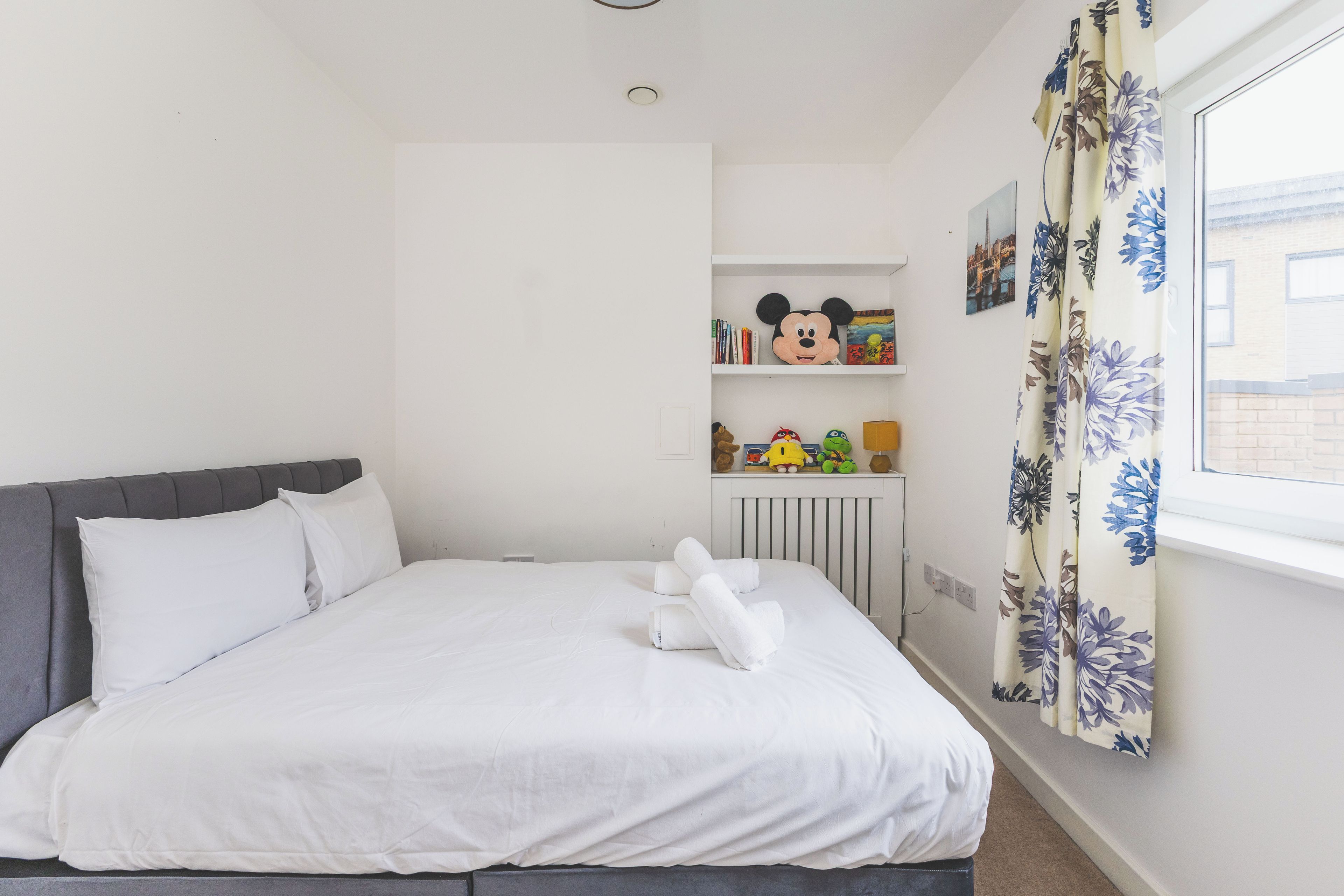 A bedroom with a bed, a radiator, a shelf with toys, and a window with curtains.