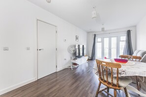 Een moderne woonkamer met een eetgedeelte, een witte deur, een staande ventilator en een groot raam met gordijnen.
