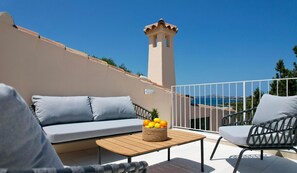 Ein Balkon mit einer Couch, einem Sessel, einem kleinen Tisch mit Früchten und Blick auf das Meer und einen Turm.